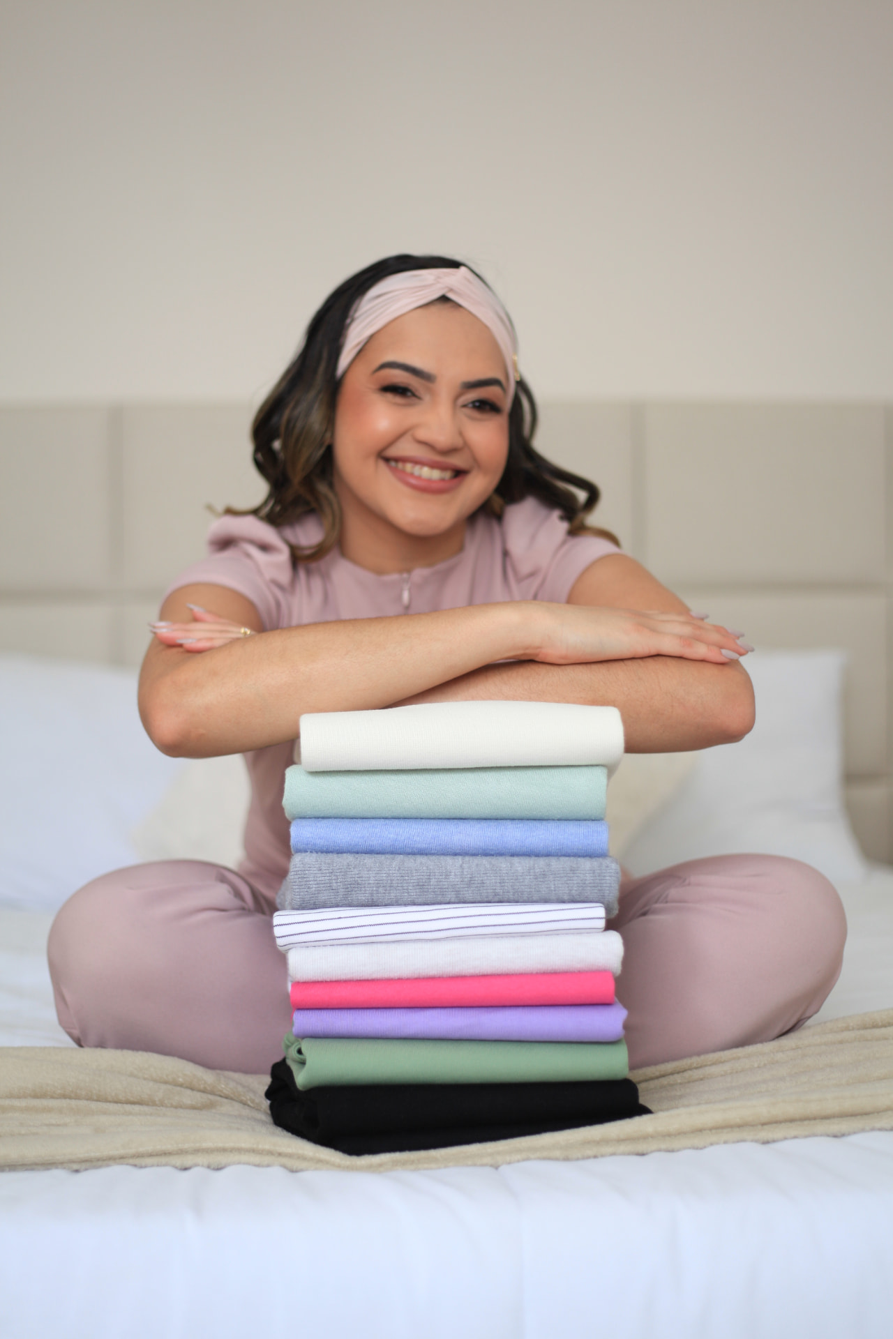 Mulher sorrindo sentada na cama com pilha de roupas coloridas